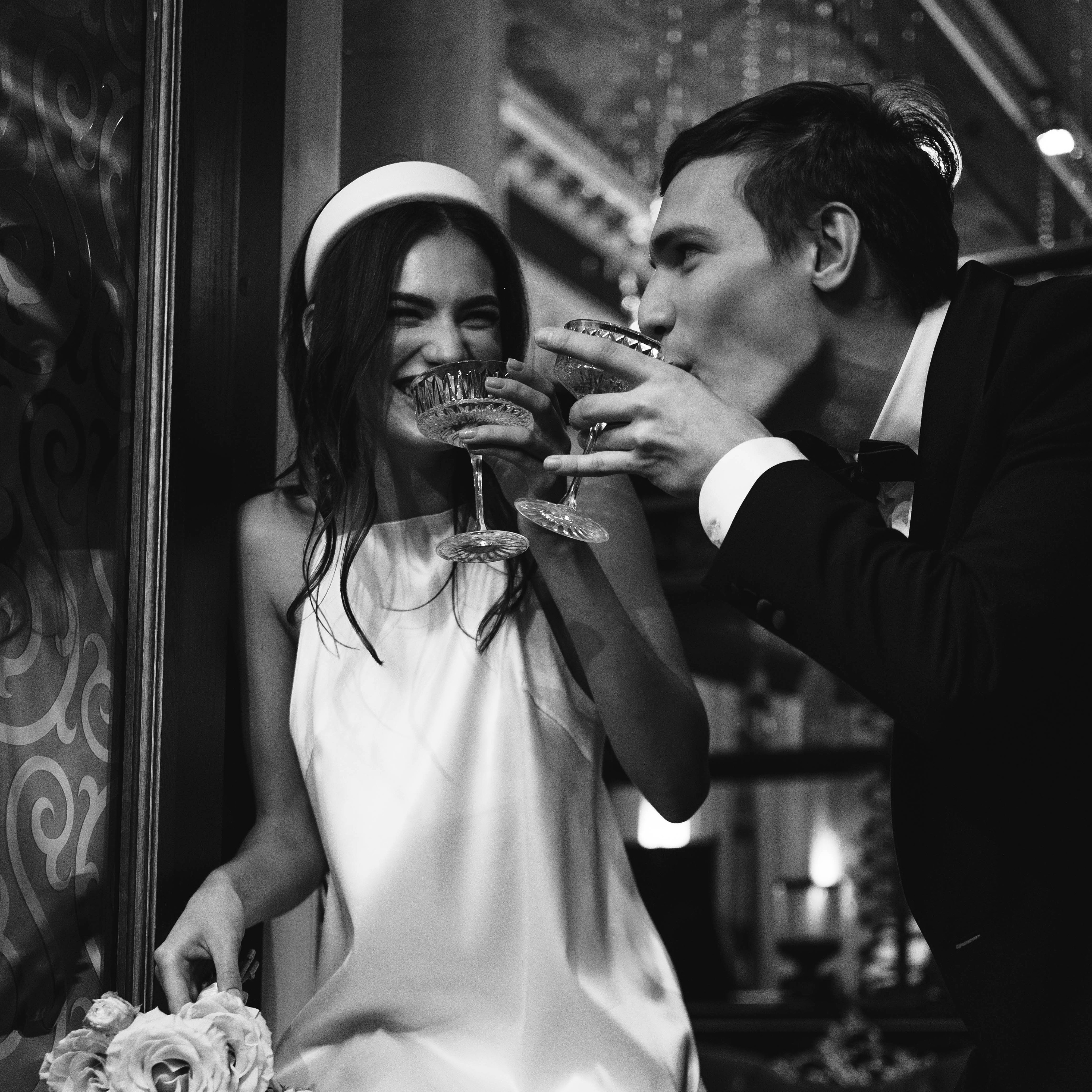 Black and white photo of a smiling couple clinking glasses and gazing into a glass of champagne, capturing a joyful and intimate moment